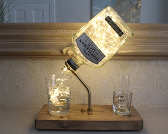May include: A repurposed Jack Daniel's Gentleman Jack whiskey bottle with a string of lights inside, mounted on a wooden base with two clear glass tumblers. The bottle is tilted to pour into one of the glasses.
