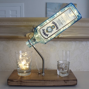 May include: A Bombay Sapphire gin bottle lamp with a blue label, filled with fairy lights, mounted on a wooden base. The lamp is connected to a metal spout and is next to two clear glasses.