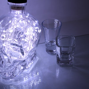 May include: A clear glass skull-shaped bottle with internal string lights, alongside two shot glasses. One shot glass features the text "Jack Daniel's Whiskey". The items are on a reflective surface.