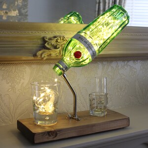 May include: A decorative lamp made from a green Tanqueray No. TEN gin bottle, with a silver spout and a wooden base. The bottle is lit with fairy lights, and the lamp is accompanied by two small glasses and a glass with fairy lights.
