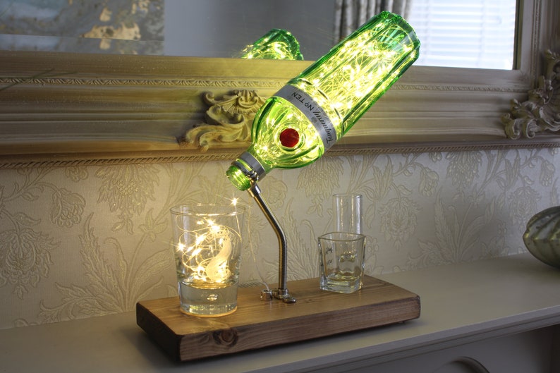 May include: A decorative lamp made from a green Tanqueray No. TEN gin bottle, with a silver spout and a wooden base. The bottle is lit with fairy lights, and the lamp is accompanied by two small glasses and a glass with fairy lights.