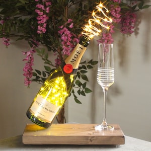 May include: A champagne bottle with a string of fairy lights wrapped around it, sitting on a wooden base. A champagne flute is next to the bottle.