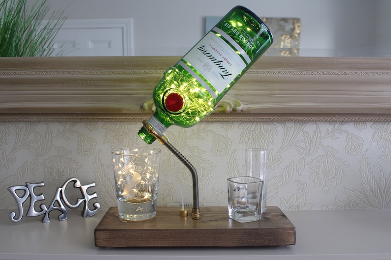 An illuminated Tanqueray gin bottle is supported in the air at a 45 degree angle by a slim arched threaded arm and brass fittings attached to a dark wooden base. The bottle is filled with 100 micro LED lights pouring out into a glass tumbler.