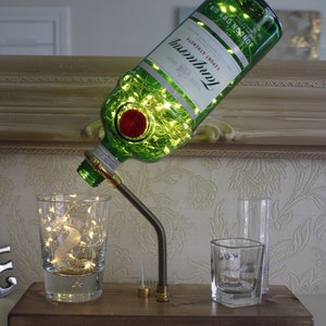 May include: A decorative lamp made from a green Tanqueray gin bottle, filled with fairy lights. The bottle is mounted on a wooden base with a metal arm, pouring into a glass. Additional glasses are also on the base.