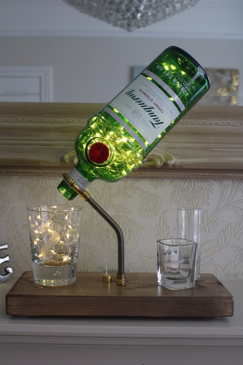May include: A decorative lamp made from a green Tanqueray gin bottle, filled with fairy lights. The bottle is mounted on a wooden base with a metal arm, pouring into a glass. Additional glasses are also on the base.