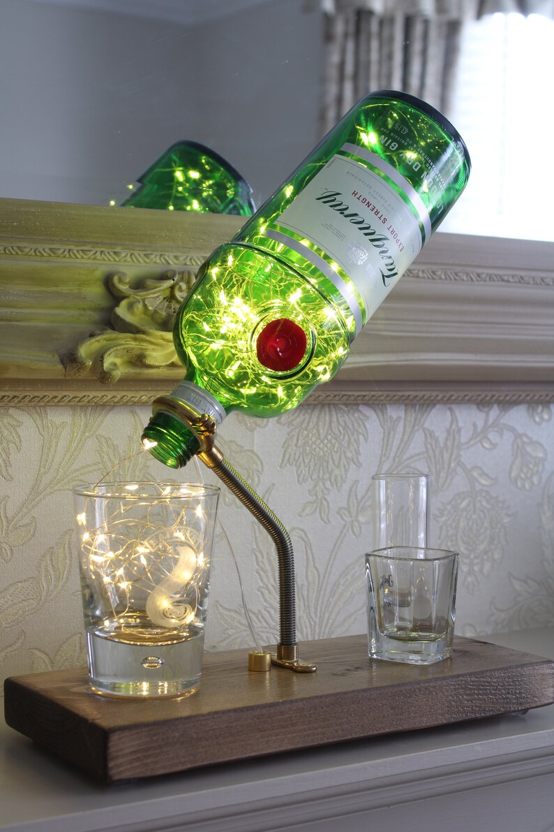 May include: A repurposed green glass bottle lamp with internal fairy lights, a red wax seal, and the Tanqueray logo. The lamp is mounted on a wooden base with a flexible metal arm, illuminating a glass and two shot glasses.