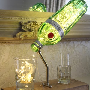 May include: A unique table lamp made from a green glass bottle, illuminated with fairy lights. The bottle is mounted on a metal gooseneck and wooden base, with two clear glasses. The bottle has a red stopper and a label that reads "Tanqueray No. TEN".