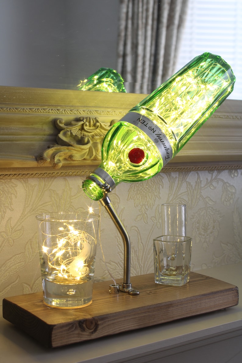 May include: A unique table lamp made from a green glass bottle, illuminated with fairy lights. The bottle is mounted on a metal gooseneck and wooden base, with two clear glasses. The bottle has a red stopper and a label that reads "Tanqueray No. TEN".