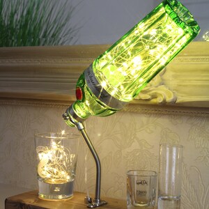 May include: A unique lamp made from a green gin bottle, filled with fairy lights, and mounted on a wooden base. The lamp is positioned above a glass with lights, and two shot glasses. The lamp's design is a creative home decor item.