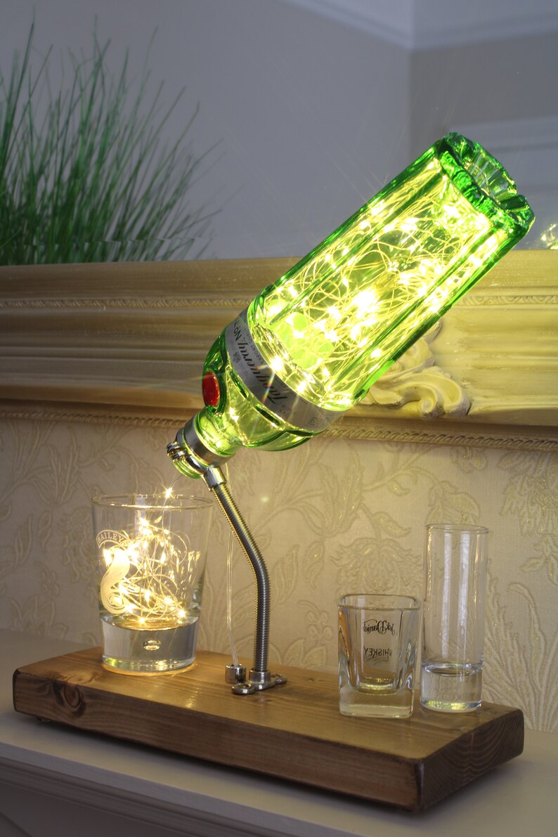 May include: A unique lamp made from a green gin bottle, filled with fairy lights, and mounted on a wooden base. The lamp is positioned above a glass with lights, and two shot glasses. The lamp's design is a creative home decor item.