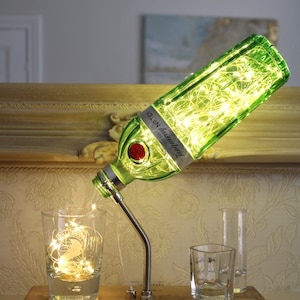May include: A repurposed gin bottle lamp with a green glass bottle filled with fairy lights. The bottle is mounted on a wooden base with a flexible metal arm. A glass with fairy lights sits below the bottle. Other glasses are on the base.