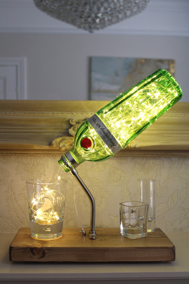 May include: A repurposed gin bottle lamp with a green glass bottle filled with fairy lights. The bottle is mounted on a wooden base with a flexible metal arm. A glass with fairy lights sits below the bottle. Other glasses are on the base.