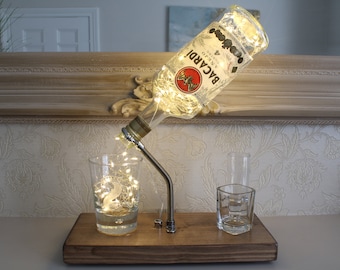 May include: A decorative lamp made from a repurposed Bacardi bottle, featuring string lights inside. The bottle is mounted on a wooden base with a metal spout, pouring into a glass. Two additional glasses are also on the base.