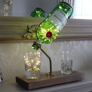 May include: A decorative lamp made from a green gin bottle with the Tanqueray label. The bottle is tilted, with fairy lights inside. The lamp is mounted on a wooden base with two small glasses. The lamp has a metal arm and a red stopper.