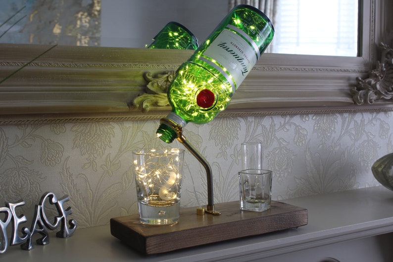 May include: A decorative lamp made from a green gin bottle with the Tanqueray label. The bottle is tilted, with fairy lights inside. The lamp is mounted on a wooden base with two small glasses. The lamp has a metal arm and a red stopper.