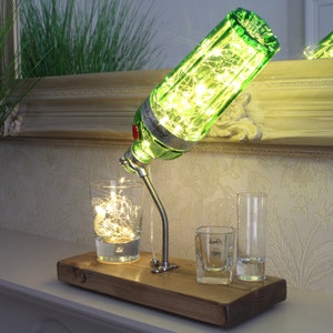 May include: A unique table lamp made from a green gin bottle, illuminated with fairy lights. The lamp is mounted on a wooden base, with three shot glasses. The lamp is on a white surface, with a mirror in the background.