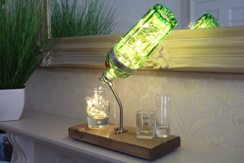 May include: A unique table lamp made from a green gin bottle, illuminated with fairy lights. The lamp is mounted on a wooden base, with three shot glasses. The lamp is on a white surface, with a mirror in the background.