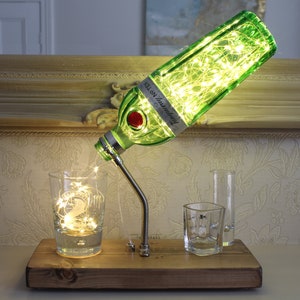 May include: A unique table lamp made from a green gin bottle, with the label "Tanqueray No. TEN". The bottle is filled with fairy lights, and the spout pours into a glass. The lamp sits on a wooden base.