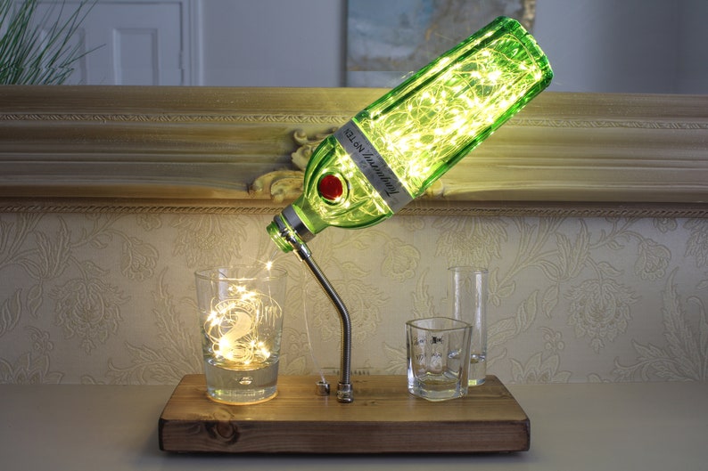 May include: A unique table lamp made from a green gin bottle, with the label "Tanqueray No. TEN". The bottle is filled with fairy lights, and the spout pours into a glass. The lamp sits on a wooden base.