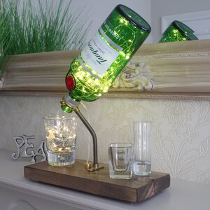 May include: A decorative lamp made from a green Tanqueray gin bottle, with string lights inside. The bottle is mounted on a wooden base with a metal spout and three shot glasses. The lamp is a unique home decor item.