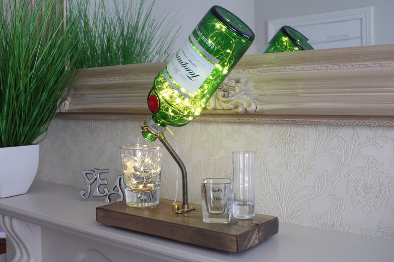 May include: A decorative lamp made from a green Tanqueray gin bottle, with string lights inside. The bottle is mounted on a wooden base with a metal spout and three shot glasses. The lamp is a unique home decor item.