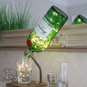 May include: A repurposed green glass bottle lamp with string lights, a metal spout, and a wooden base. The lamp is accompanied by three clear shot glasses, one of which has text. The bottle is labeled "Tanqueray London Dry".