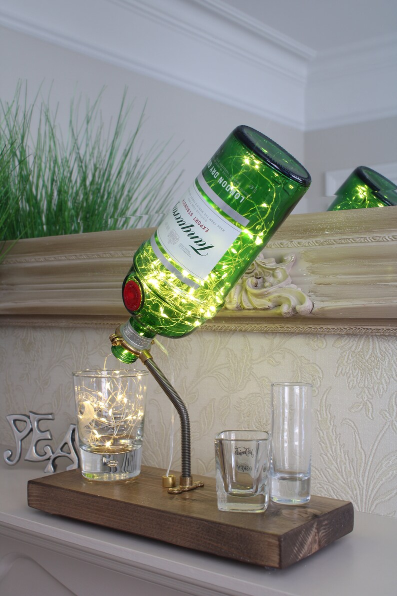 May include: A repurposed green glass bottle lamp with string lights, a metal spout, and a wooden base. The lamp is accompanied by three clear shot glasses, one of which has text. The bottle is labeled "Tanqueray London Dry".