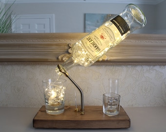 May include: A repurposed liquor bottle lamp with the label "OAKHEART BACARDI CARTA" and string lights inside. The bottle is mounted on a wooden base with a metal spout, pouring into a glass. Two additional glasses are on the base.
