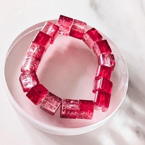 May include: A red and clear glass bracelet made of square beads. The bracelet is on a white marble surface.