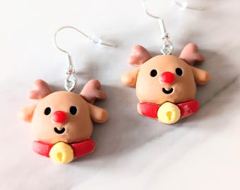 May include: A pair of Christmas-themed earrings featuring reindeer designs. Each earring has a light brown reindeer with a red nose, small antlers, and a red scarf with a yellow bell. The earrings hang from silver-colored hooks.