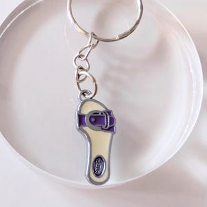 May include: A silver key chain with a cream-colored sandal charm. The sandal has a purple strap with a buckle and the word "Crocs" printed on the sole.
