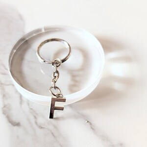 May include: A silver-coloured keychain with the letter "F" pendant. The keychain is attached to a small chain and a circular key ring. The background is a white, marble-patterned surface.