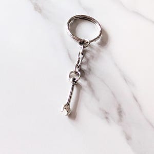 May include: A silver wrench-shaped keychain. The keychain features a small, detailed wrench charm suspended from a chain and a keyring. The background is a white marble surface.
