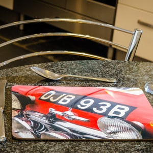 Op de afbeelding: Rode auto met een zwart kenteken waarop "DJB 93B" staat. De auto staat op een bruine onderzetter met een zwarte achtergrond.