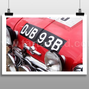 Op de afbeelding: Close-up van een rode vintage auto met een zwart-wit kenteken waarop "DJB 93B" staat. De auto heeft meerdere koplampen en een chromen bumper.