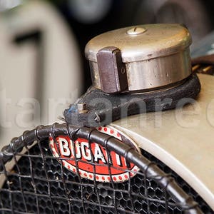 Op de afbeelding: Close-up van een vintage Bugatti-auto met een zilveren tankdop en een zwart rooster met het woord "BUGATTI" in rode letters.