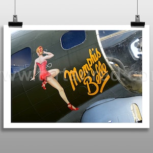 Könnte beinhalten: Ein grünes und graues Bomberflugzeug aus dem Zweiten Weltkrieg mit den Worten "Memphis Belle" in Gelb an der Seite. Ein Pin-up-Girl in einem roten Kleid ist an der Seite des Flugzeugs gemalt.