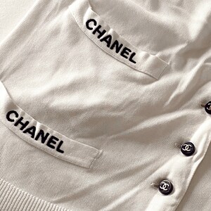 May include: White cardigan with black embroidered "CHANEL" on the pockets and black button closures with the Chanel logo.