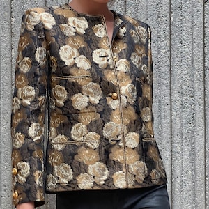 Op de afbeelding: Een zwart en gouden jas met een bloemenpatroon en gouden knopen. De jas heeft een mandarijnkraag en lange mouwen.