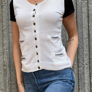 May include: White short-sleeved cardigan with black sleeves and a button-up front. The cardigan has the word "CHANEL" embroidered on the front and back.