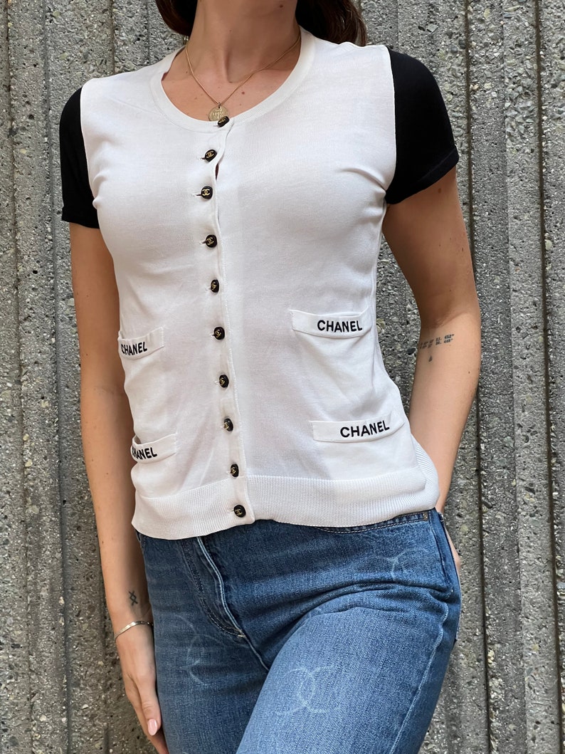 May include: White short-sleeved cardigan with black sleeves and a button-up front. The cardigan has the word "CHANEL" embroidered on the front and back.