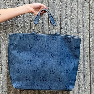 May include: A blue denim tote bag with a clear plastic chain handle and a repeating pattern of the Chanel logo.