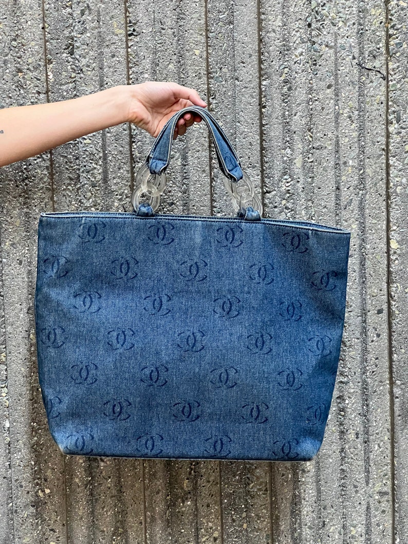 May include: A blue denim tote bag with a clear plastic chain handle and a repeating pattern of the Chanel logo.
