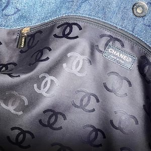 May include: The inside of a blue denim bag is lined with black fabric featuring a repeating pattern of the Chanel logo. A black leather tag with the Chanel logo and "Made in Italy" text is visible.