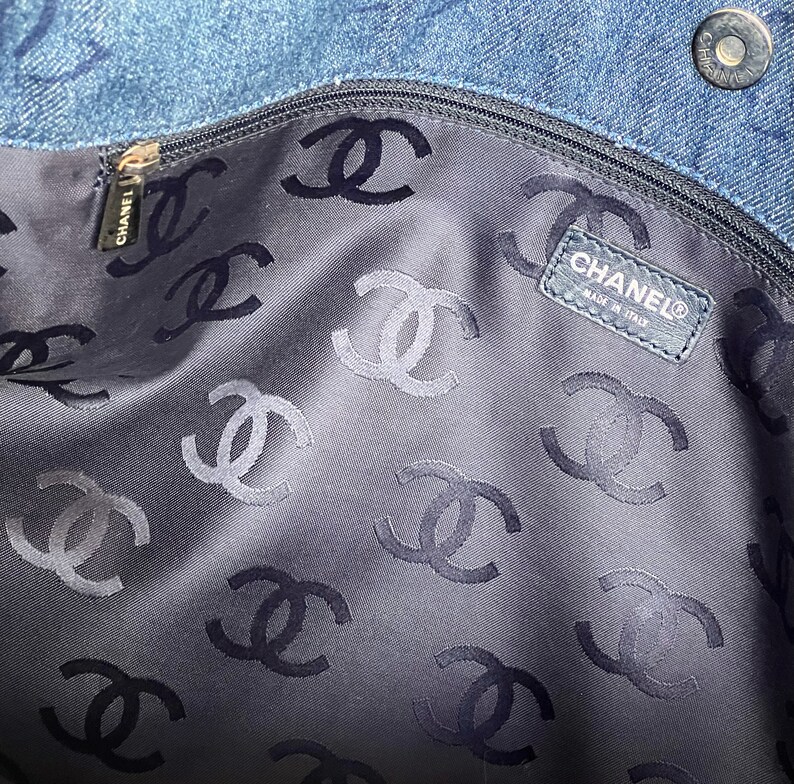 May include: The inside of a blue denim bag is lined with black fabric featuring a repeating pattern of the Chanel logo. A black leather tag with the Chanel logo and "Made in Italy" text is visible.