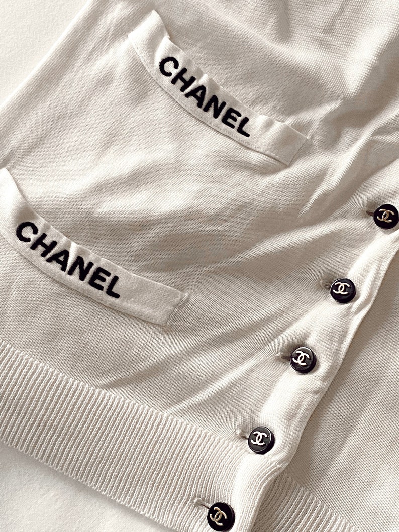 May include: White cardigan with black embroidered "CHANEL" on the pockets and black button closures with the Chanel logo.
