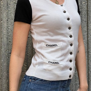 May include: White short-sleeved cardigan with black sleeves and a button-up front. The cardigan has two pockets with the word "CHANEL" embroidered on them.