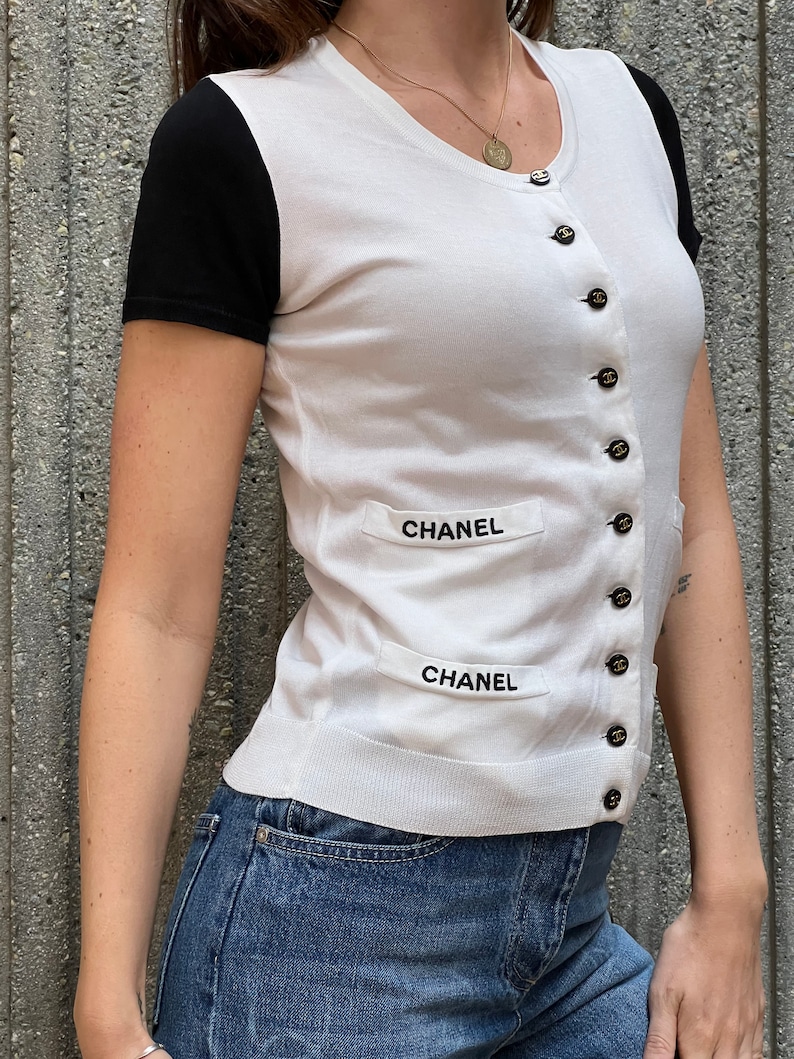 May include: White short-sleeved cardigan with black sleeves and a button-up front. The cardigan has two pockets with the word "CHANEL" embroidered on them.