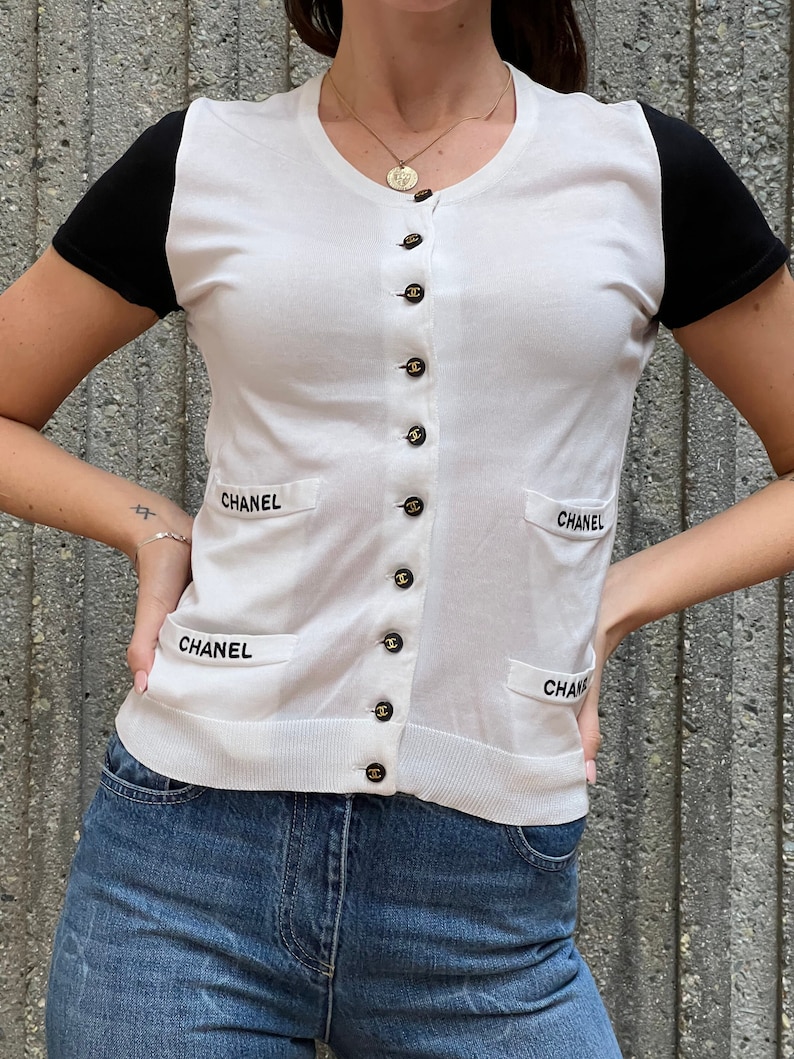 May include: White short-sleeved cardigan with black sleeves and a button-up front. The cardigan has the word "CHANEL" embroidered on the front and back.