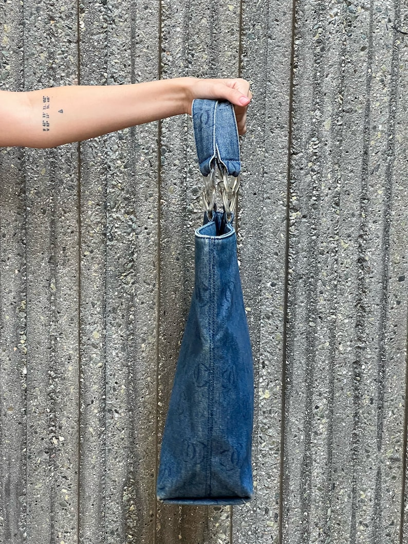 May include: A blue denim tote bag with a silver chain strap. The bag has a faded look and is made of a durable material.
